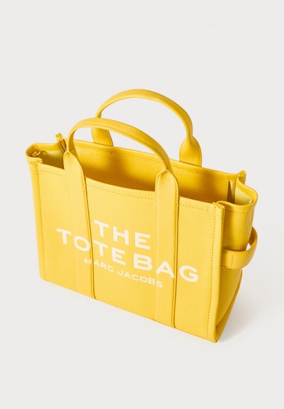 Yellow leather tote bag with double handles, side pocket, and white text reading "THE TOTE BAG MARC JACOBS." Textured finish and structured shape.