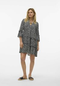 Vestido negro con estampado floral, de mangas largas, con accentos de volantes y escote en V. Tela ligera con un diseño en capas. Llevado con sandalias deslizables en color marino.
