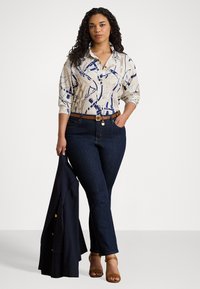 Camicia a maniche lunghe con bottoni di colore crema con un motivo astratto blu, abbinata a jeans di un blu scuro e una cintura marrone. Sandali con tacco marroni.