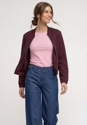 Ung kvinde smiler, iført en lys rosa skjorte, åben mørkerød jakke og højtaljede løse blå jeans mod en ensfarvet baggrund.