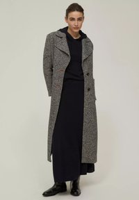 Cappotto grigio a scacchi con colletto, due bottoni e tasche laterali. Indossato sopra un hoodie nero e un lungo vestito. Stivaletti neri alla caviglia completano il look.