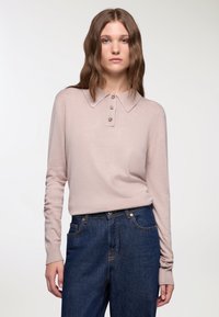 Kobieta z falującymi brązowymi włosami, ubrana w jasnoró�żową, długą koszulę polo i ciemnoniebieskie dżinsy z wysokim stanem, na białym tle.