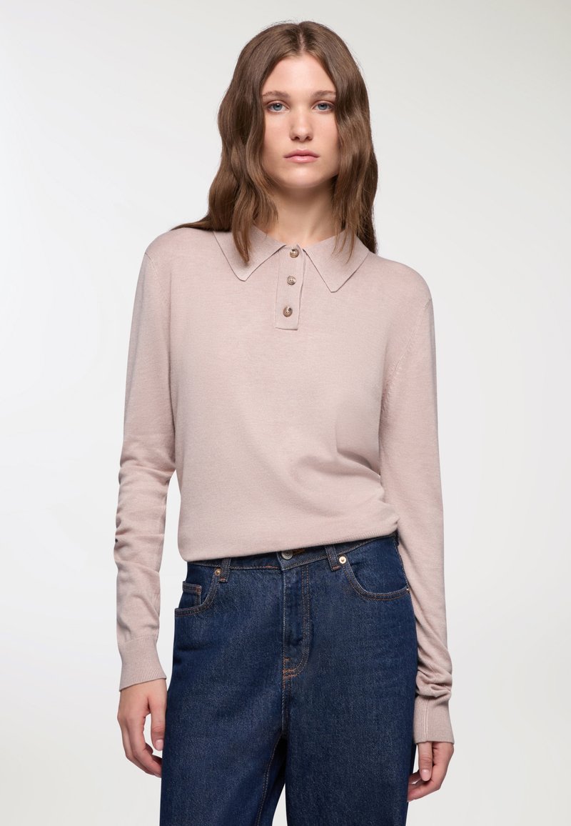 Kobieta z falującymi brązowymi włosami, ubrana w jasnoróżową, długą koszulę polo i ciemnoniebieskie dżinsy z wysokim stanem, na białym tle.