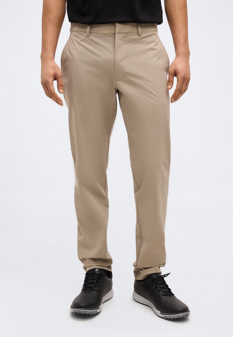 Άνδρας που φοράει μπεζ παντελόνι ίσιας γραμμής και μαύρα δετά παπούτσια, με λευκό φόντο.