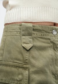 Pantaloni cargo verde măsliniu cu finisaj texturat, prevăzuți cu închidere cu nasture și o buclă pentru curea înclinată distinctivă în talie.