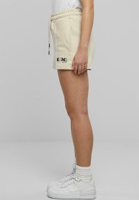 Beige bomullsshorts med elastisk midja, snörning, två sidofickor och färggård "KAN" broderi på vänstra låret.
