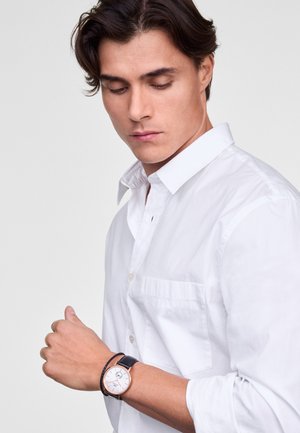 Round, analog watch with a white face, black leather strap, and rose gold accents. Worn on a wrist, featuring a button-up white shirt.