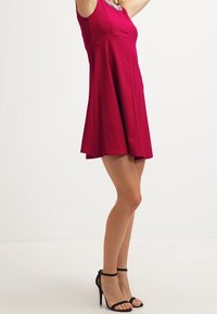 Fuchsia ärmelloses Kleid mit ausgestelltem Rock und rundem Ausschnitt, das eine strukturierte Verzierung am Hals hat. Schwarze Riemchen-Pumps.