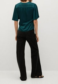 Blouse en satin bleu sarcelle avec manches courtes évasées et dos plissé, associée à un jean noir à jambes larges et des sandales plates.