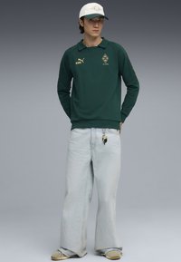 Tânăr îmbrăcat cu un hanorac dark green Puma cu logo-uri, blugi largi de culoare deschisă cu chei atârnanți, adidași bej și o șapcă de baseball albă.