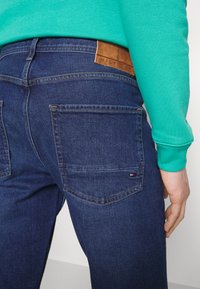 I jeans in denim blu scuro presentano tasche a toppa e un'etichetta in pelle marrone sul cinturino. Il maglione è turchese con polsini a coste.