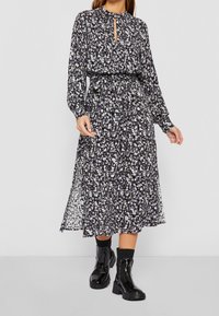 Langärmliges, floral gemustertes Kleid in dunklen Farbtönen mit einem Schlüssellochausschnitt und tailliertem Bund, kombiniert mit schwarzen Knöchelschuhen.
