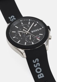 Black chronograph watch with a stainless steel case, textured black dial, white hour markers, and silicone strap featuring "BOSS" in gray.