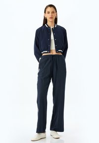 Marineblauwe cropped bomberjack met witte gestreepte manchetten en kraag, gecombineerd met losse marineblauwe broeken met zijstrepen, gedragen met lichte sneakers.