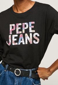 Svart långärmad t-shirt med ett färgglatt blommönster på "PEPE JEANS"-logotypen; ihop med ett denimbälte och högmidjade jeans.