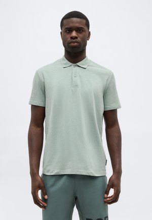 Noormees seisab, seljas heleroheline lühikeste varrukatega polo särk ja tumerohelised lühikesed püksid, ilma erilise taustata valgel taustal.