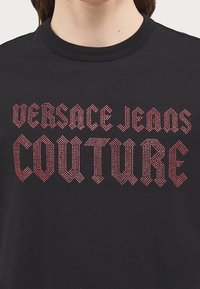 Černé bavlněné tričko s potiskem "VERSACE JEANS COUTURE" z vyvýšených červených kamínků. Kulatý výstřih, krátké rukávy a přiléhavý střih.
