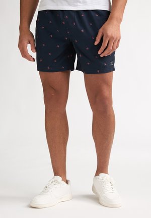 Shorts de bain bleu marine avec un motif d'ancre rose subtil, taille élastique et poches latérales. Portés avec des baskets blanches.