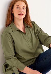 Olivgrön, avslappnad skjorta utan krage med sidofickor och trekvartslånga ärmar, i kombination med mörk denimbyxor.