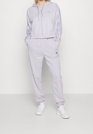 Kobieta ubrana w jasnofioletowy dres Adidas z bluzą z kapturem i zamkiem, w paski na rękawach, pasujące spodnie oraz białe trampki, stojąca z rękami w kieszeniach.