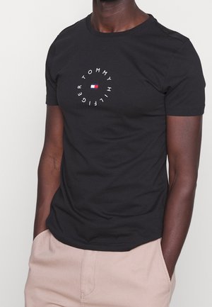 Mand iført sort kortærmet T-shirt med cirkulær "Tommy Hilfiger" tekst og logo, parret med beige bukser, hænderne i lommerne.