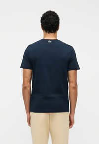 Άνδρας με σγουρά μαλλιά που φοράει μπλε ναυτικό μπλουζάκι με μικρό λογότυπο στο πάνω μέρος της πλά της και μπεζ παντελόνι, γυρισμένος με την πλάτη στην κάμερα.