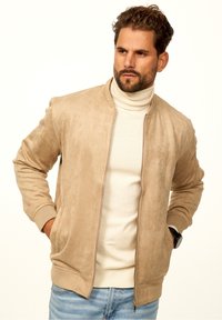 Beige suede bomber jacket with a zip front, ribbed cuffs, and two side pockets, layered over a cream turtleneck sweater.