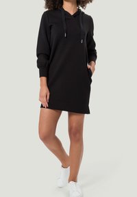 Robe sweat à capuche noire avec manches longues, poches latérales et cordons ajustables. Texture lisse avec une coupe décontractée, se terminant au-dessus du milieu de la cuisse.