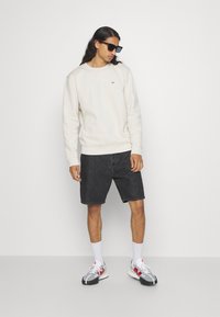 Cremefarbener Pullover, dunkle Jeansshorts und graue Sportschuhe mit roten Akzenten. Das Model trägt Sonnenbrillen und steht vor einem neutralen Hintergrund.