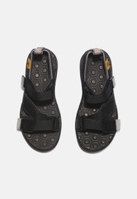 Chinelos pretos com duas correias ajustáveis, sola de borracha texturizada com padrão hexagonal e uma etiqueta de logo perto do dedo do pé.