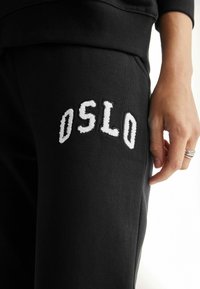 Crne trenirke s izvezenim "OSLO" logotipom u bijeloj boji na lijevoj natkoljenici, izrađene od mekanog materijala s opuštenim krojem i bočnim džepovima.