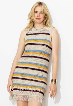 Femme blonde portant une robe sans manches en crochet avec des rayures horizontales crème, bleues, jaunes et bordeaux et un ourlet à franges, debout devant un fond blanc.