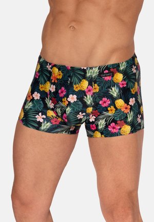 Torso masculin portant un short de bain noir avec un imprimé tropical coloré de fleurs et d'ananas, portant la marque "olaf benz" sur la ceinture.