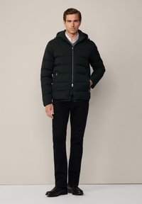 Uomo che indossa un giubbotto imbottito nero con cappuccio, un maglione grigio, una camicia bianca, pantaloni neri e scarpe nere, in piedi davanti a uno sfondo semplice.