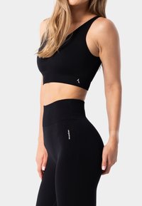 Top corto atlético negro con una tirante en el hombro, combinado con leggings de cintura alta negros que presentan el logo de la marca en blanco en la pierna. Textura suave.