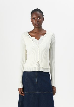 Hvit ribbet cardigan med lange ermer, med v-hals og knappelukking. Bæres over en grå topp og kombinert med et denimskjørt.