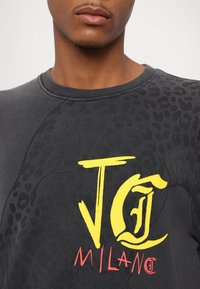 Grå hettegenser med en teksturert svart leopardprint og en dristig gul "JG" grafikk, akkompagnert av rød "MILANO" tekst. Myk stoff, rund halslinning.
