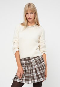 Pull en maille crème à manches retroussées, porté avec une jupe plissée à carreaux dans des tons bruns et blancs. Modèle portant des collants foncés.