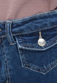 Σκούρα μπλε τζιν με μπροστινή τσέπη, ασημένιο κουμπί και αντίθετη ραφή κατά μήκος της μέσης και των ακρών της τσέπης.