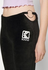 Crne velur hlače s naglaskom u obliku srebrnog prstena na džepu i izvezenim "KANI" logom. Tkanina ima mekanu teksturu.