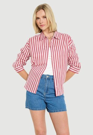 Blonde Frau, die ein rot-weiß gestreiftes Hemd mit Knöpfen über einem weißen Oberteil und blauen Jeansshorts trägt, steht mit hinter dem Rücken verschränkten Händen.