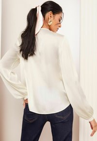 Weiße Bluse mit langen Ärmeln aus strukturiertem Stoff, mit elastischen Bündchen und abgerundetem Saum. Kombiniert mit dunklen Jeans und einem Haartuch.