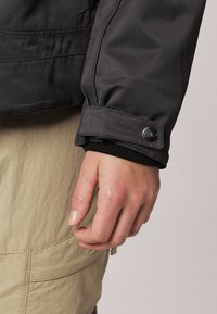 Gros plan d’une main reposant près d’une jambe de pantalon beige à poche, portant une manche de veste foncée avec un poignet boutonné.