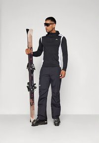 Zwart-wit ski-outfit met een gevoerde capuchontrui met rits, bijpassende broek en zwarte skischoenen; houdt bruine en zwarte ski's vast.