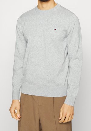 Mand iført lys grå langærmet bluse med rund hals og lille rød, hvid og blå logo på brystet samt brune bukser.