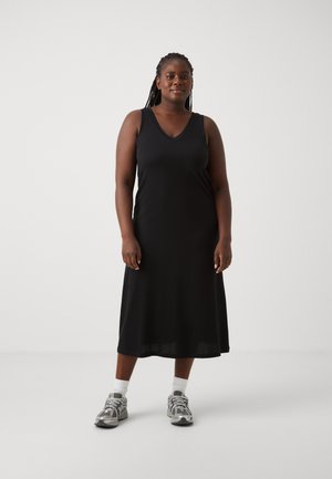 Robe noire sans manches avec un décolleté en V, faite d'un tissu lisse, tombant mi-mollet. Associée à des chaussures de sport grises et des chaussettes blanches.