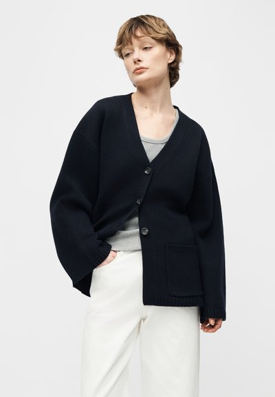 Άτομο που φοράει ένα φαρδύ μαύρο πλεκτό ζακέτα με κουμπιά και μπροστινή τσέπη πάνω από γκρι πουκάμισο και λευκό παντελόνι, στέκεται μπροστά σε απλό φόντο.