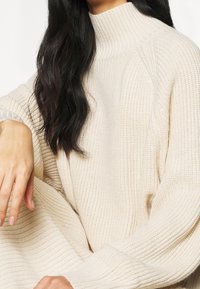 Maglione a collo alto lavorato a maglia color crema con texture a coste, maniche ampie e vestibilità rilassata, che mette in evidenza un design accogliente adatto per il layering.
