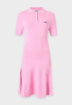 Femme portant une robe polo rose, des chaussettes roses, des talons noirs et portant un petit sac noir à bandoulière, debout devant un fond blanc uni.