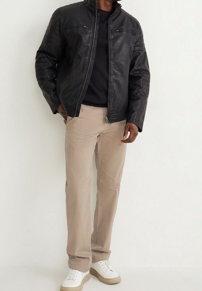 Homme portant une veste en cuir noire, une chemise noire, un pantalon beige et des baskets blanches, debout devant un fond blanc uni.